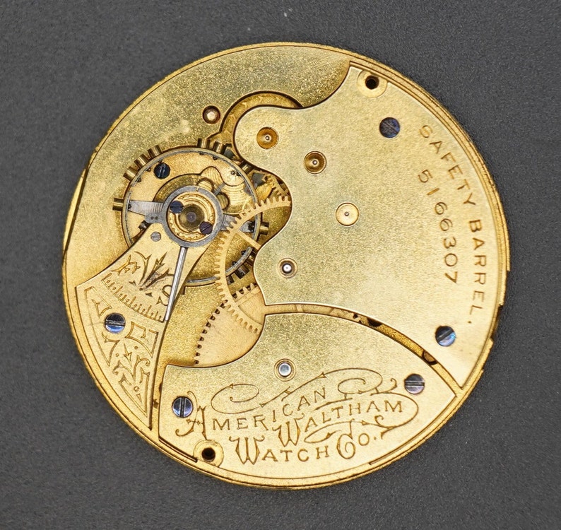 Puede incluir: Primer plano de un movimiento de reloj de bolsillo de color dorado con la inscripci&oacute;n "SAFETY BARREL 5166307" y "AMERICAN WALTHAM WATCH CO."