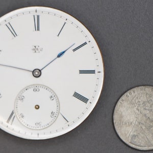 May include: A white pocket watch face with black Roman numeral hour markers and black minute markers. The watch has a blue second hand and a small white sub-dial for seconds. The watch is about the size of a US quarter.
