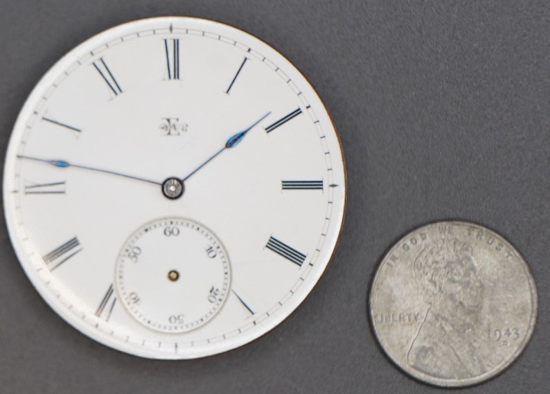 May include: A white pocket watch face with black Roman numeral hour markers and black minute markers. The watch has a blue second hand and a small white sub-dial for seconds. The watch is about the size of a US quarter.