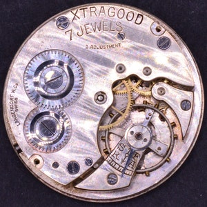 Pu&ograve; includere: Primo piano di un movimento di orologio da tasca argentato con il testo "Xtragood 7 Jewels 1 Adjustment" e "Langendorf W.Co. Swiss" sul quadrante.