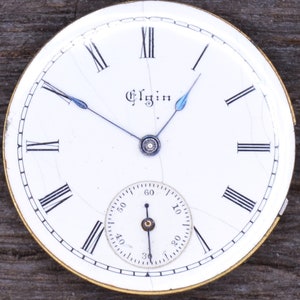 May include: White pocket watch face with black Roman numeral hour markers and black minute markers. The watch face has a small, white sub-dial with black markings. The watch face is cracked and has a gold-coloured rim. The watch face says "Elgin" in the centre.