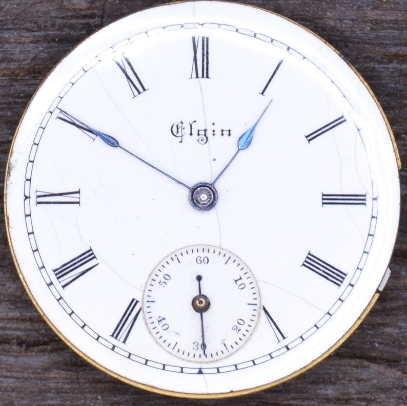 May include: White pocket watch face with black Roman numeral hour markers and black minute markers. The watch face has a small, white sub-dial with black markings. The watch face is cracked and has a gold-coloured rim. The watch face says "Elgin" in the centre.