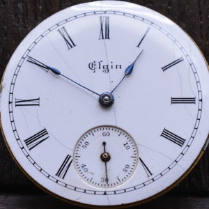 May include: A white pocket watch face with black Roman numeral hour markers and black minute markers. The watch face has a small, white sub-dial with black markings for seconds. The watch face is signed "Elgin" in black lettering.