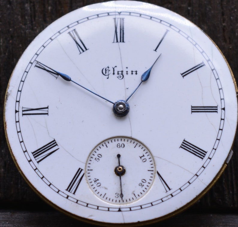 May include: A white pocket watch face with black Roman numeral hour markers and black minute markers. The watch face has a small, white sub-dial with black markings for seconds. The watch face is signed "Elgin" in black lettering.