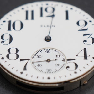 Op de afbeelding: Een wit zakhorloge met zwarte Romeinse cijfers als uurmarkeringen en zwarte wijzers. De wijzerplaat van het horloge bevat ook een kleinere, ronde sub-wijzerplaat met zwarte markeringen en wijzers. Het horloge is bovenaan met "ELGIN" gelabeld.