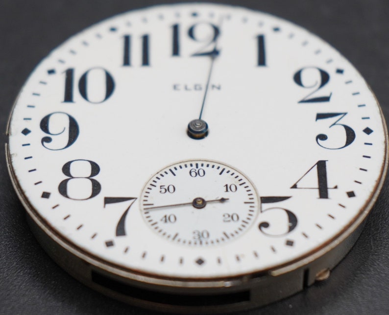 Op de afbeelding: Een wit zakhorloge met zwarte Romeinse cijfers als uurmarkeringen en zwarte wijzers. De wijzerplaat van het horloge bevat ook een kleinere, ronde sub-wijzerplaat met zwarte markeringen en wijzers. Het horloge is bovenaan met "ELGIN" gelabeld.
