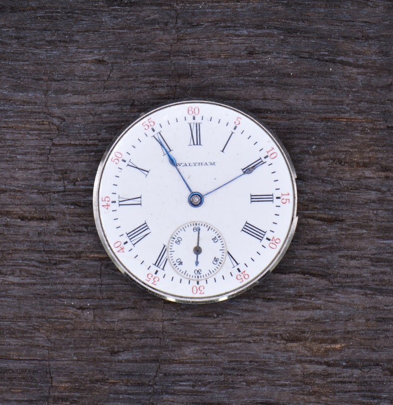 May include: A white pocket watch with Roman numeral markings and a blue hand. The watch face has the word "WALTHAM" printed on it.