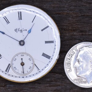 May include: A white pocket watch face with black Roman numeral hour markers and black minute markers. The watch face is cracked and has a small, white sub-dial with black markings. The watch face is inscribed with the word "Elgin". The watch face is about the size of a UK ten pence coin.