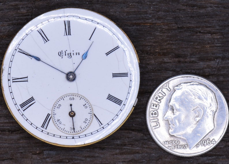 May include: A white pocket watch face with black Roman numeral hour markers and black minute markers. The watch face is cracked and has a small, white sub-dial with black markings. The watch face is inscribed with the word "Elgin". The watch face is about the size of a UK ten pence coin.