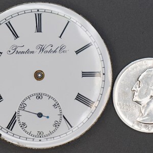 May include: White pocket watch face with black Roman numeral hour markers and black minute markers. The watch face has the text "Trenton Watch Co." printed on it. The watch face is about the size of a US quarter.