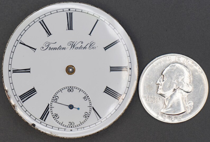 May include: White pocket watch face with black Roman numeral hour markers and black minute markers. The watch face has the text "Trenton Watch Co." printed on it. The watch face is about the size of a US quarter.