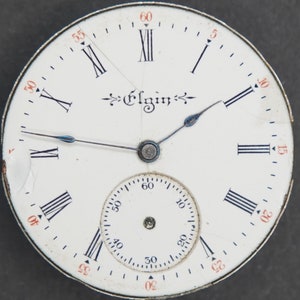 Puede incluir: Una esfera de reloj de bolsillo blanca con marcadores de hora en n&uacute;meros romanos negros y marcadores de minutos negros. La esfera del reloj tiene un c&iacute;rculo conc&eacute;ntrico m&aacute;s peque&ntilde;o con marcadores de minutos negros y la palabra "Elgin" impresa en negro.
