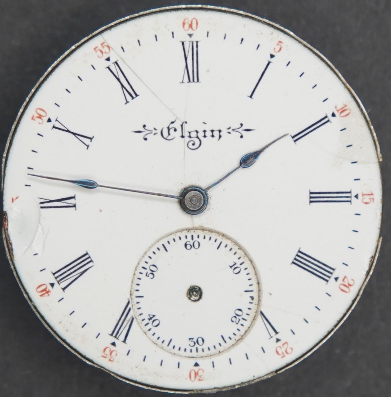 Puede incluir: Una esfera de reloj de bolsillo blanca con marcadores de hora en n&uacute;meros romanos negros y marcadores de minutos negros. La esfera del reloj tiene un c&iacute;rculo conc&eacute;ntrico m&aacute;s peque&ntilde;o con marcadores de minutos negros y la palabra "Elgin" impresa en negro.