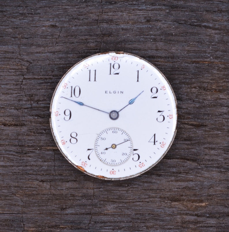 Puede incluir: Un reloj de bolsillo blanco con agujas negras y un peque&ntilde;o subdial. La esfera del reloj tiene n&uacute;meros negros y la palabra "ELGIN" impresa en ella.