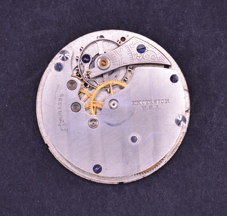 May include: Close-up of the back of a silver pocket watch with a gold-colored gear system. The watch is labeled "EXCELSIOR U.S.A." and has the number "4274058285" printed on the back.