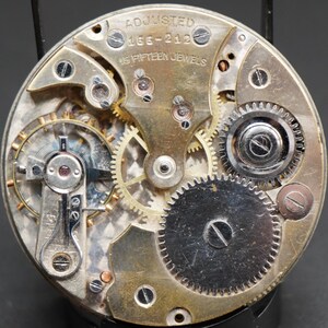 May include: Close-up of a silver and gold watch movement with the text "ADJUSTED 166-212 15 FIFTEEN JEWELS".