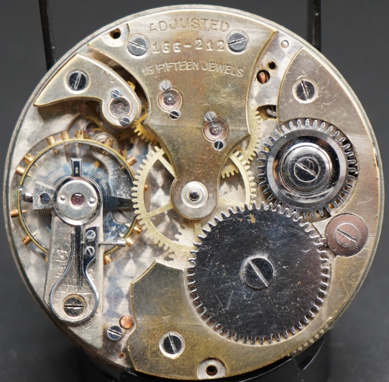 May include: Close-up of a silver and gold watch movement with the text "ADJUSTED 166-212 15 FIFTEEN JEWELS".
