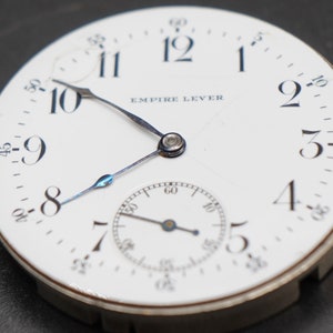 May include: A white pocket watch with black Roman numeral hour markers and black Arabic numeral minute markers. The watch face has the text "EMPIRE LEVER" printed on it. The watch has a small second hand dial at the bottom.