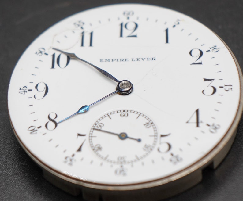 May include: A white pocket watch with black Roman numeral hour markers and black Arabic numeral minute markers. The watch face has the text "EMPIRE LEVER" printed on it. The watch has a small second hand dial at the bottom.