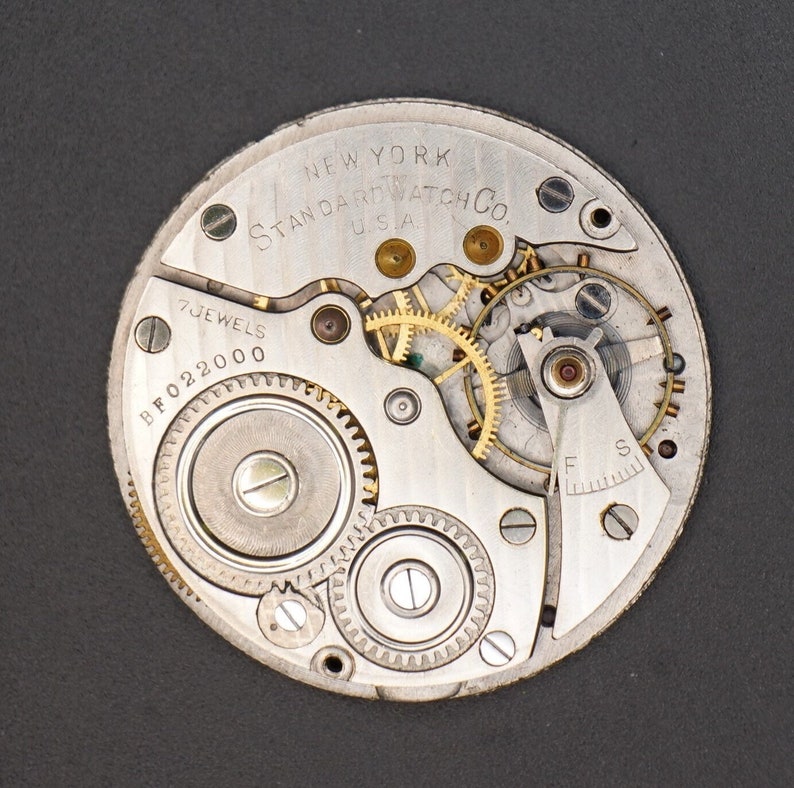 May include: Close-up view of a silver pocket watch movement with gold gears. The watch face reads "NEW YORK STANDARD WATCH CO. USA" and "7 JEWELS BF022000".