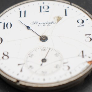 May include: A white pocket watch with black Roman numeral hour markers and a black second hand. The watch face is cracked and has the text "Standard U.S.A." printed on it.