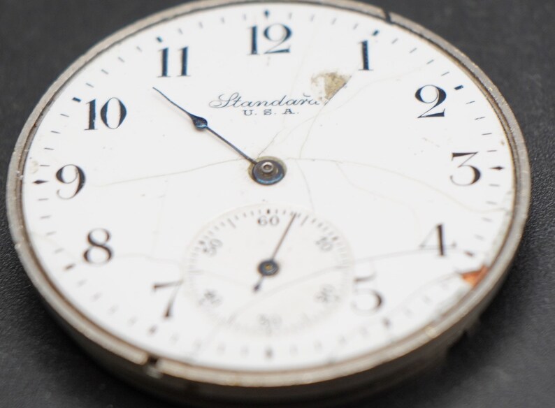 May include: A white pocket watch with black Roman numeral hour markers and a black second hand. The watch face is cracked and has the text "Standard U.S.A." printed on it.