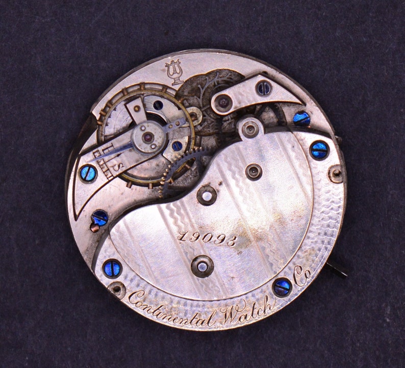 May include: Close-up of the back of a silver pocket watch with a detailed mechanical movement. The watch face has the inscription "Continental Watch Co." and the number "19093".