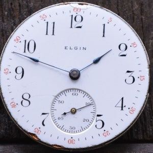 Puede incluir: Un reloj de bolsillo blanco con marcadores de hora romanos negros y agujas negras. La esfera del reloj tiene la palabra "ELGIN" impresa en negro. El reloj tiene un subdial blanco m&aacute;s peque&ntilde;o con marcas negras.