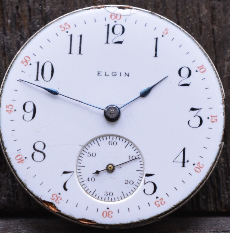 Puede incluir: Un reloj de bolsillo blanco con marcadores de hora romanos negros y agujas negras. La esfera del reloj tiene la palabra "ELGIN" impresa en negro. El reloj tiene un subdial blanco m&aacute;s peque&ntilde;o con marcas negras.