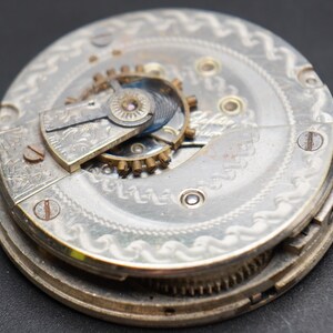 May include: Close-up of a silver pocket watch movement with intricate engravings and gears. The watch face is visible in the background.