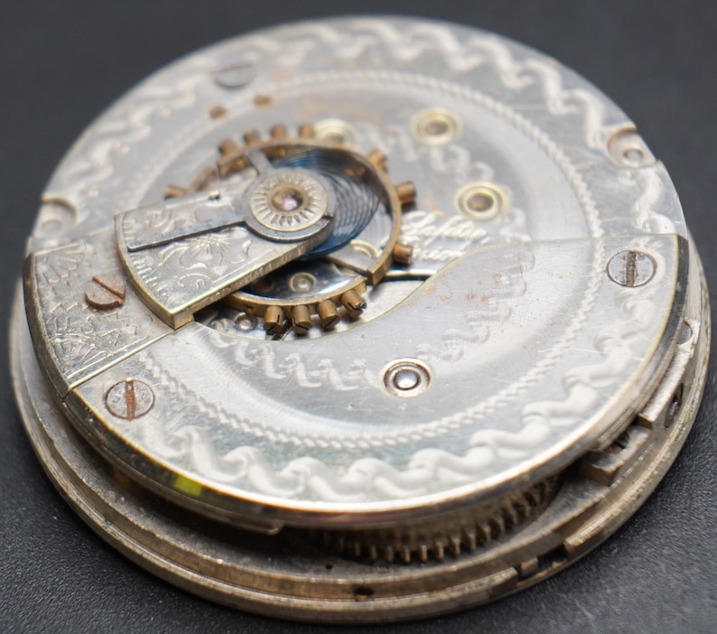May include: Close-up of a silver pocket watch movement with intricate engravings and gears. The watch face is visible in the background.