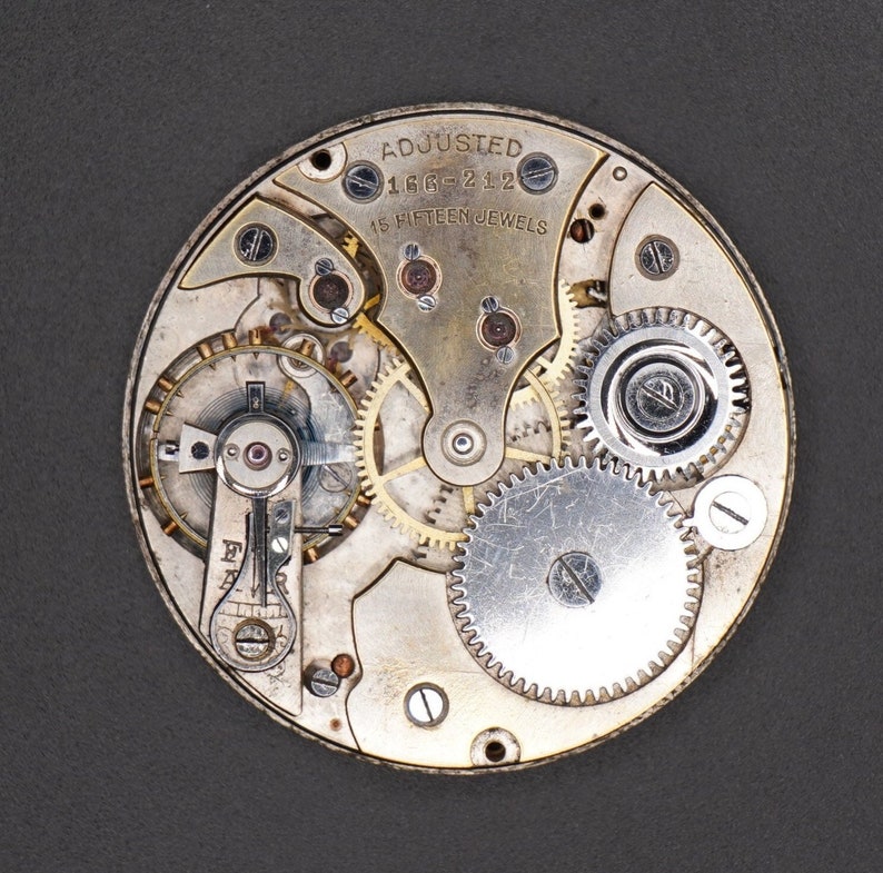 May include: Close-up of a silver pocket watch movement with intricate gears and the text "Adjusted 166-212 15 Fifteen Jewels".
