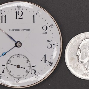 May include: A white pocket watch with black Roman numeral hour markers and black hands. The watch face reads "Empire Lever". The watch is next to a silver US quarter.