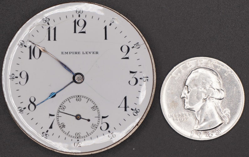 May include: A white pocket watch with black Roman numeral hour markers and black hands. The watch face reads "Empire Lever". The watch is next to a silver US quarter.