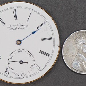 Puede incluir: Una esfera de reloj de bolsillo blanca con marcadores de hora en n&uacute;meros romanos negros y marcadores de minutos negros. La esfera del reloj tiene un peque&ntilde;o subdial blanco con marcadores de minutos negros. La esfera del reloj est&aacute; inscrita con "American Waltham". La esfera del reloj est&aacute; ligeramente da&ntilde;ada en el lado derecho. La esfera del reloj est&aacute; junto a una moneda de diez centavos estadounidense de plata.