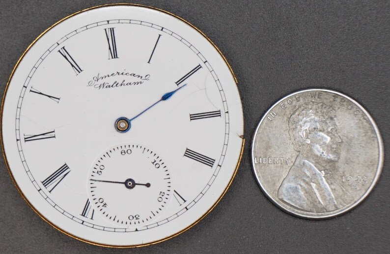Puede incluir: Una esfera de reloj de bolsillo blanca con marcadores de hora en n&uacute;meros romanos negros y marcadores de minutos negros. La esfera del reloj tiene un peque&ntilde;o subdial blanco con marcadores de minutos negros. La esfera del reloj est&aacute; inscrita con "American Waltham". La esfera del reloj est&aacute; ligeramente da&ntilde;ada en el lado derecho. La esfera del reloj est&aacute; junto a una moneda de diez centavos estadounidense de plata.