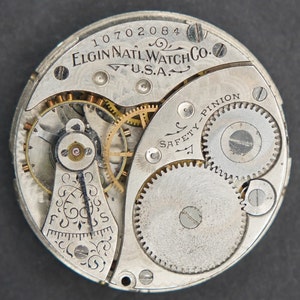 Puede incluir: Primer plano de un movimiento de reloj de bolsillo plateado con el texto "ELGIN NAT'L WATCH CO. U.S.A." y "SAFETY PINION" visible. El movimiento del reloj tiene una variedad de engranajes y resortes.