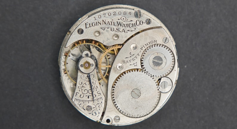 Puede incluir: Primer plano de un movimiento de reloj de bolsillo plateado con el texto "ELGIN NAT'L WATCH CO. U.S.A." y "SAFETY PINION" visible. El movimiento del reloj tiene una variedad de engranajes y resortes.
