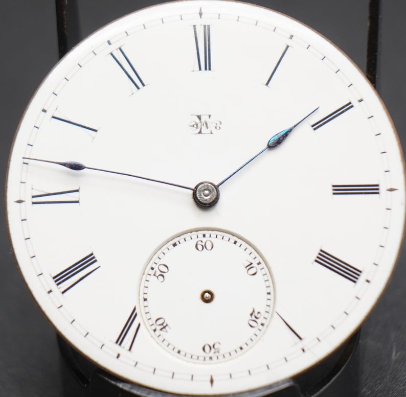 May include: A white pocket watch face with black Roman numeral hour markers and black minute markers. The watch has a small, white sub-dial with black numbers for seconds. The watch face has a blue steel hand for the seconds and a black steel hand for the minutes. The watch face has the letter "E" with a number "3" above it.