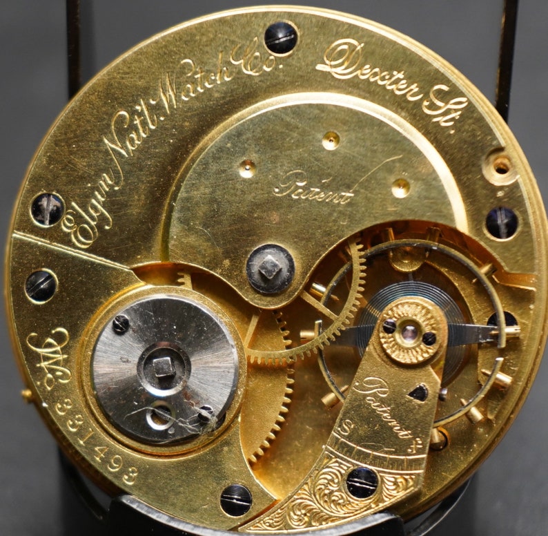 May include: Close-up of a gold-toned pocket watch movement with intricate gears and the inscription "Elgin Natl. Watch Co. Dexter St. Patent" visible.