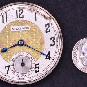 Pu&ograve; includere: Un orologio da tasca d'argento con un quadrante bianco e indici orari romani neri. L'orologio ha un quadrante interno color oro con un motivo a rombi e la scritta "STRATFORD" stampata su di esso. L'orologio &egrave; accanto a una moneta da dieci centesimi americana d'argento.