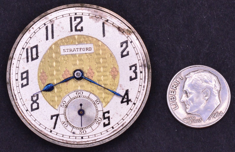 Pu&ograve; includere: Un orologio da tasca d'argento con un quadrante bianco e indici orari romani neri. L'orologio ha un quadrante interno color oro con un motivo a rombi e la scritta "STRATFORD" stampata su di esso. L'orologio &egrave; accanto a una moneta da dieci centesimi americana d'argento.