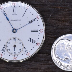May include: A white pocket watch with Roman numeral hour markers and a smaller sub-dial. The watch face says "Waltham" and has blue hands. The watch is next to a silver US dime with the year 1964.