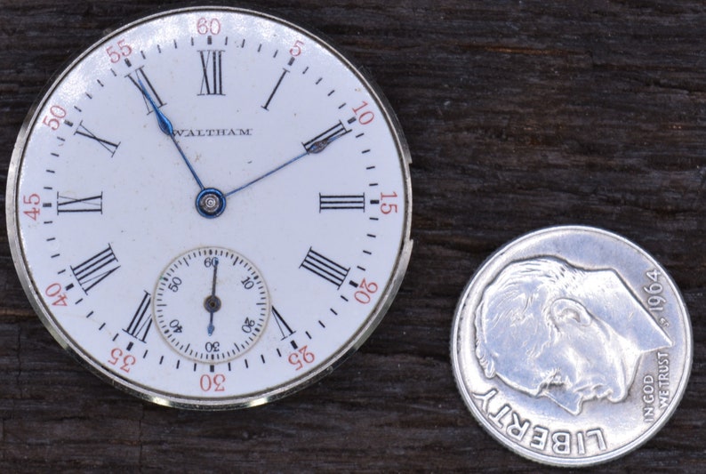 May include: A white pocket watch with Roman numeral hour markers and a smaller sub-dial. The watch face says "Waltham" and has blue hands. The watch is next to a silver US dime with the year 1964.