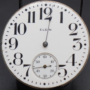 Op de afbeelding: Een wit zakhorloge met zwarte Romeinse cijfers voor de uren en zwarte wijzers. Het horloge heeft de tekst "ELGIN" op de wijzerplaat. Het horloge heeft een kleinere, ronde wijzerplaat met zwarte wijzers en zwarte markeringen voor seconden, minuten en uren.