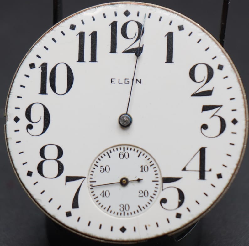 Op de afbeelding: Een wit zakhorloge met zwarte Romeinse cijfers voor de uren en zwarte wijzers. Het horloge heeft de tekst "ELGIN" op de wijzerplaat. Het horloge heeft een kleinere, ronde wijzerplaat met zwarte wijzers en zwarte markeringen voor seconden, minuten en uren.