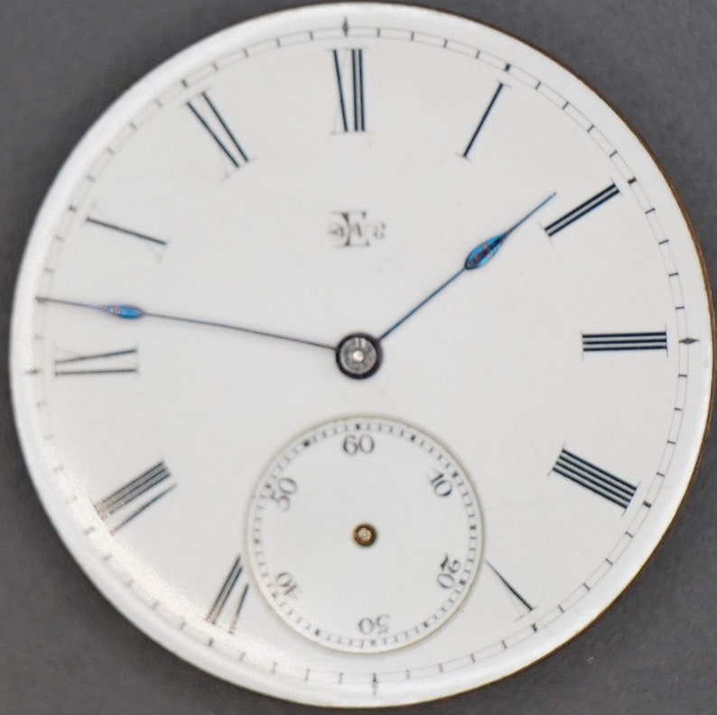 May include: White pocket watch face with black Roman numeral hour markers and black minute markers. The watch has a smaller white sub-dial with black minute markers and black numbers for the seconds. The watch hands are black with blue tips.