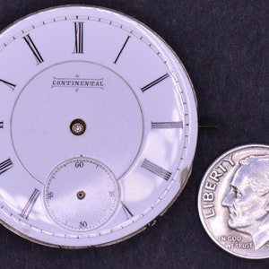 May include: White pocket watch face with black Roman numeral hour markers and black minute markers. The watch face has the text "CONTINENTAL" printed on it. The watch face is about the size of a US dime.