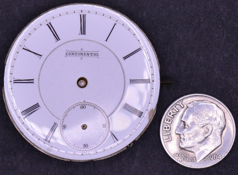 May include: White pocket watch face with black Roman numeral hour markers and black minute markers. The watch face has the text "CONTINENTAL" printed on it. The watch face is about the size of a US dime.