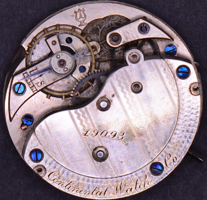 May include: Close-up of the back of a silver pocket watch with a blue steel screw design. The watch face has the text "Continental Watch Co." and the number "19093".